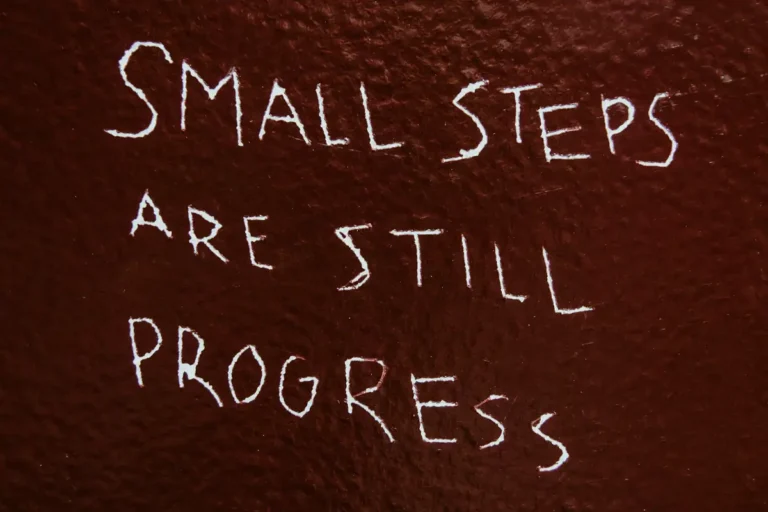 Chalk handwriting on a dark red wall reading “Small steps are still progress”