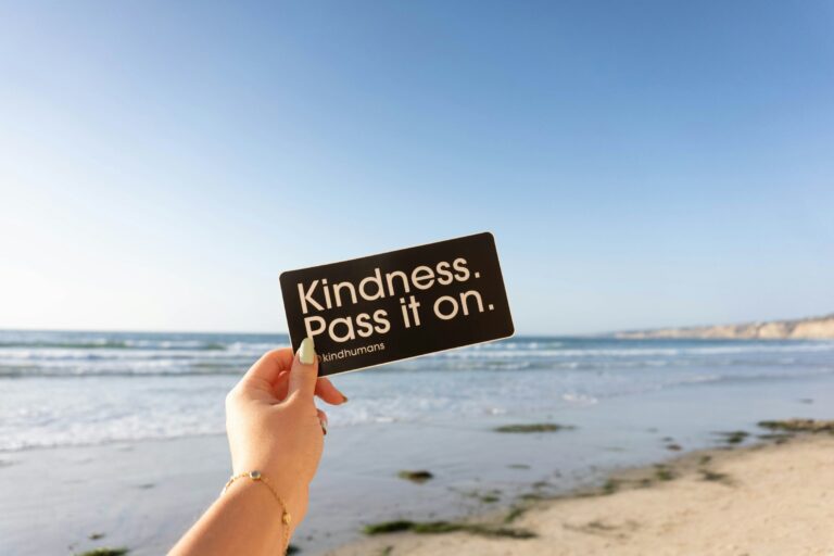 A hand holding a black card at the beach that reads "Kindness. Pass it on."