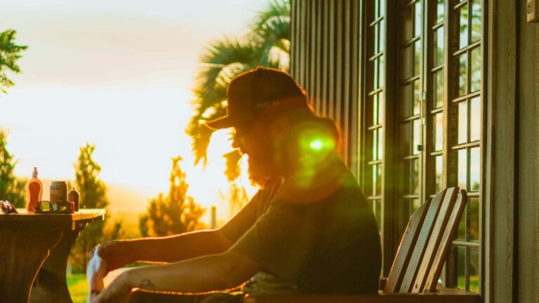A man sitting alone on a wooden porch chair at sunset, the low golden light behind him