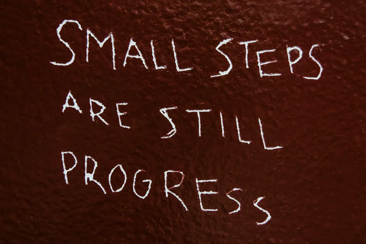 Chalk handwriting on a dark red wall reading “Small steps are still progress”